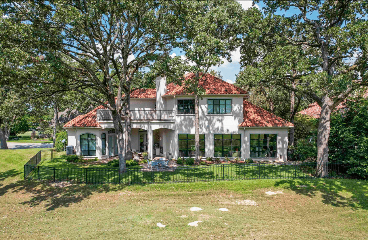 Exterior rear view of a two-story luxury home with terra cotta tile roof, arched entryways, upper balcony, and black iron fencing, surrounded by mature oak trees in The Cascades neighborhood, Tyler, TX