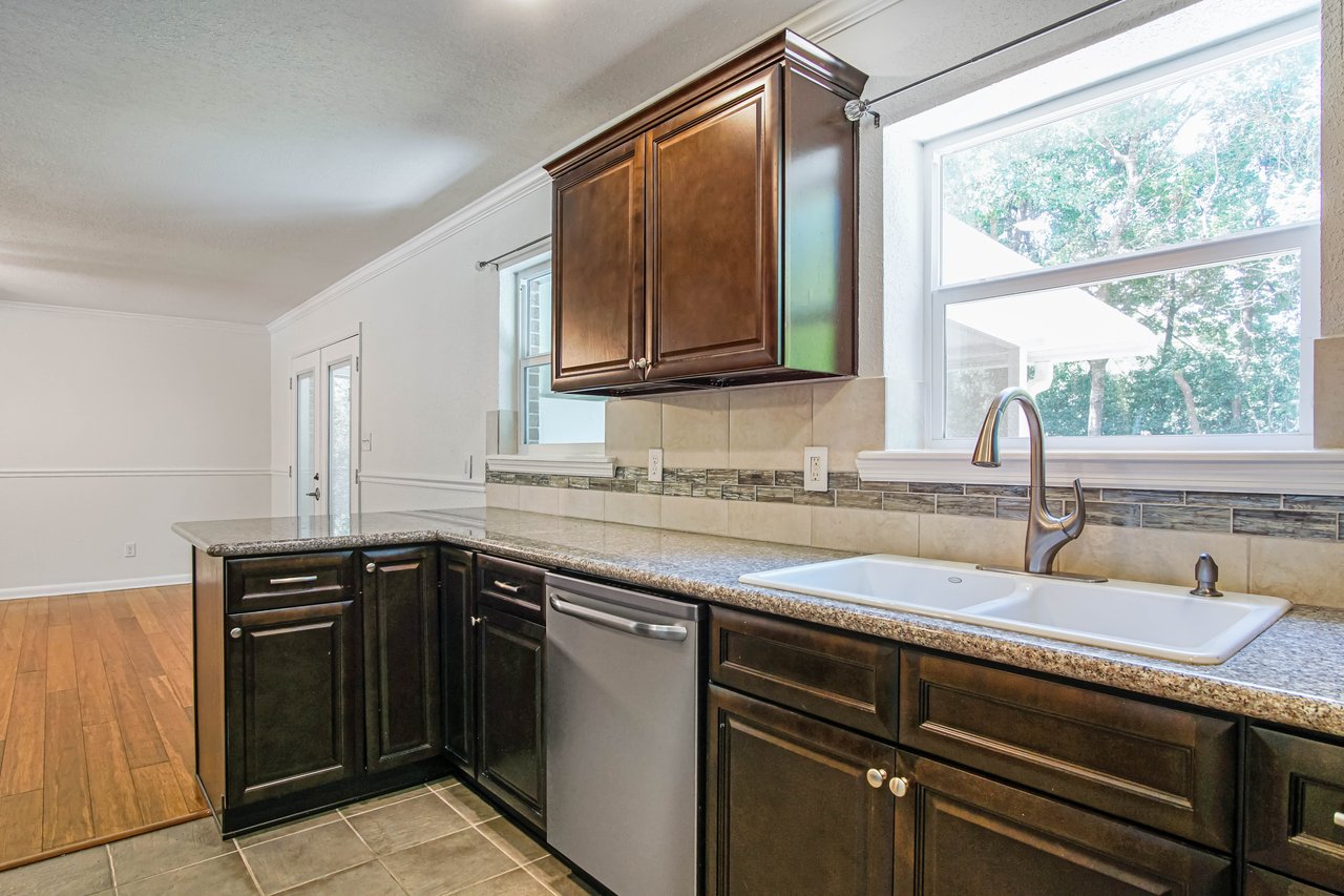 Modern kitchen with dark wood cabinets and granite countertops. A large window above a white sink, letting in natural light. Calm, clean atmosphere.