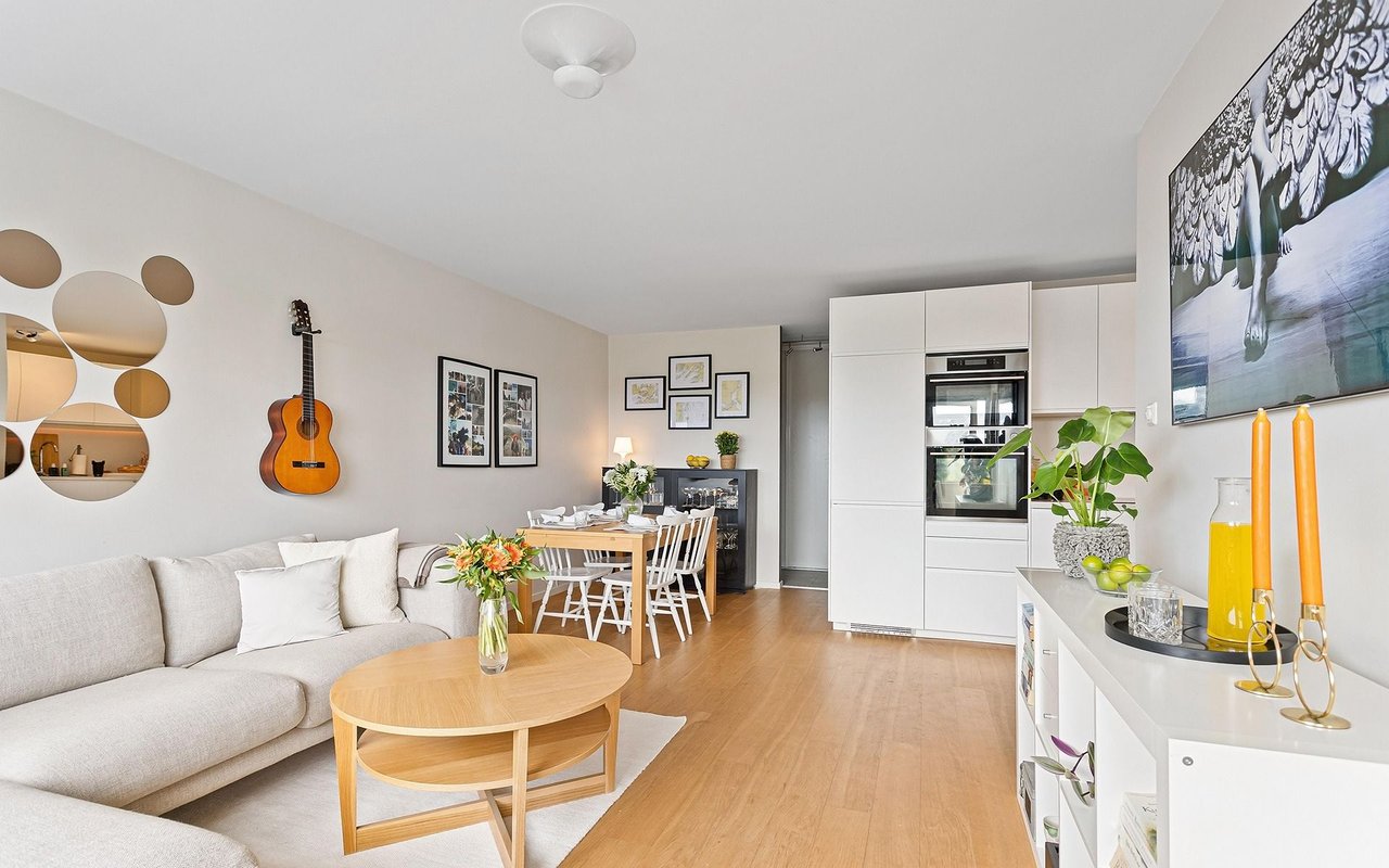 Open-plan living room with a white sofa, light wood coffee table, dining area, and modern kitchen.