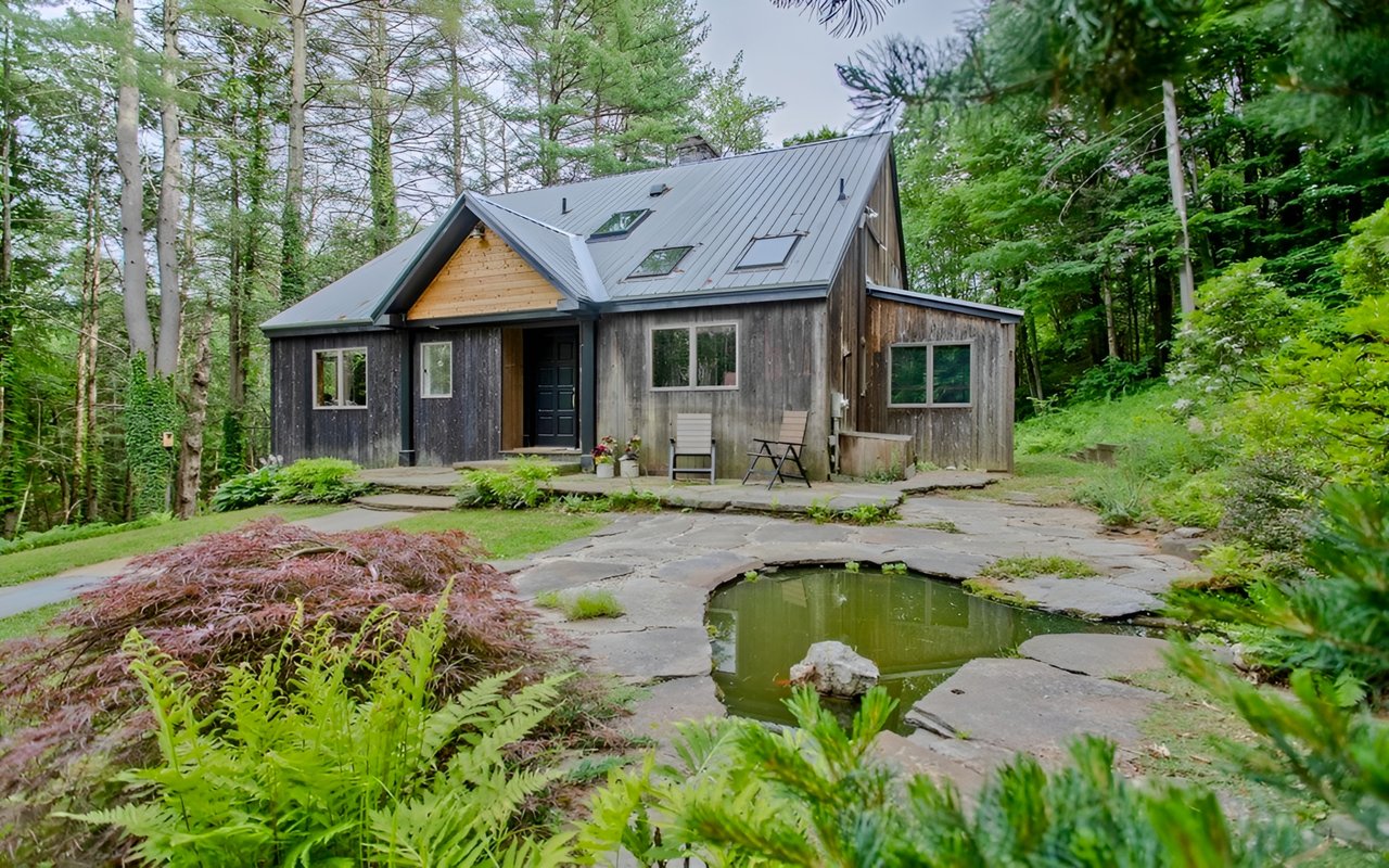 Wood-sided home among trees, with a metal roof, stone patio, and small pond.