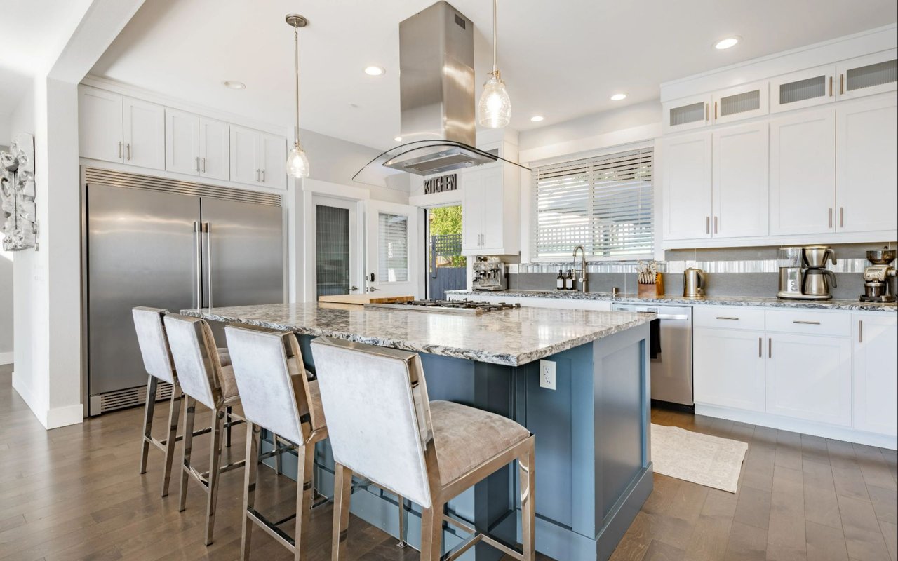 Bright white kitchen with stainless appliances, a large blue island, patterned stone countertop, and upholstered bar stools.