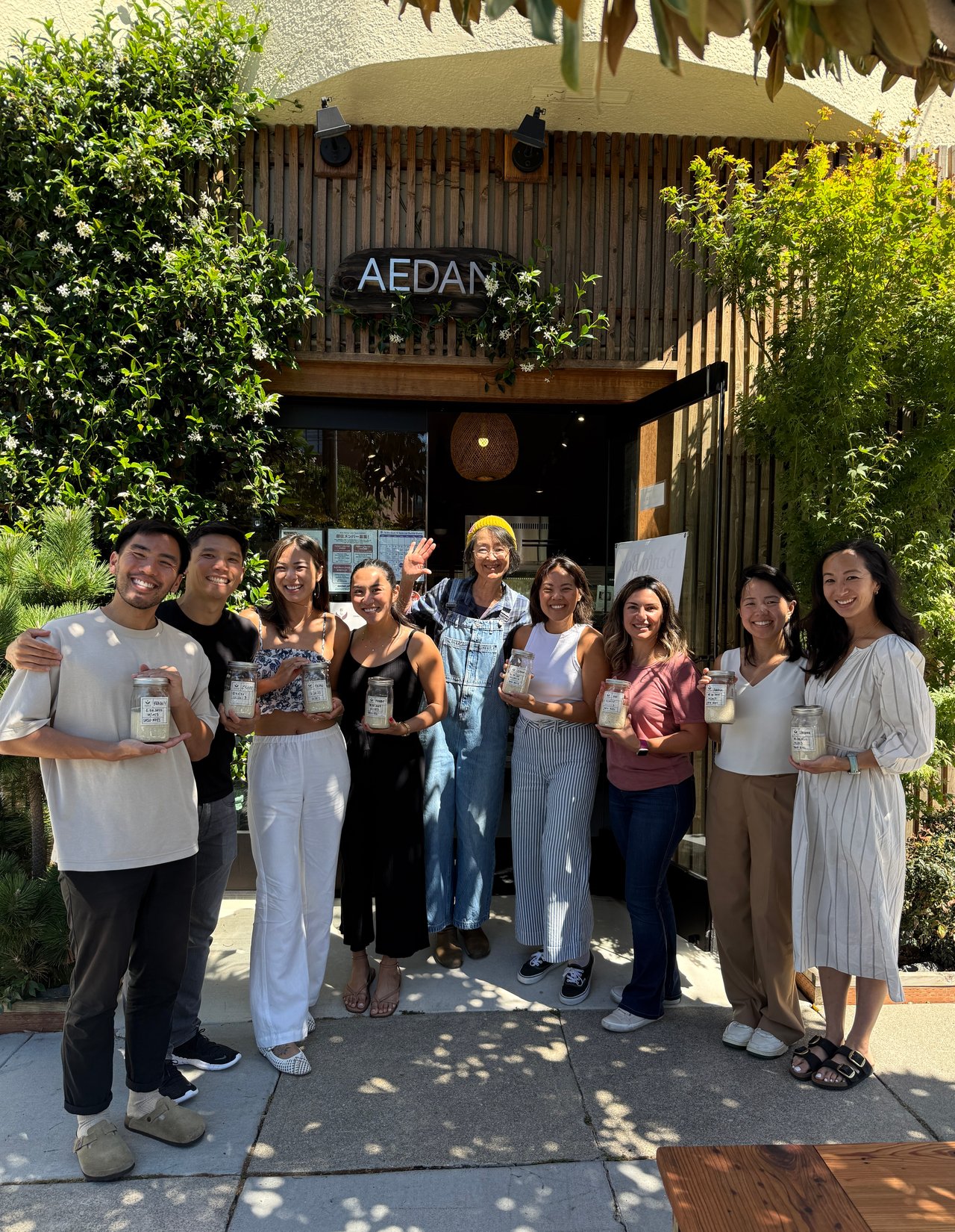 Nine adults smile and pose with jars outside a leafy storefront marked AEDAN.