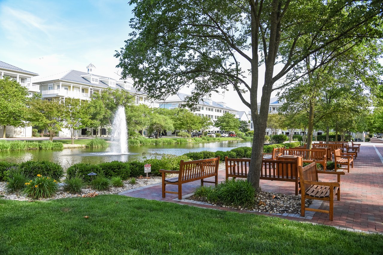Park pond and fountain beside a brick path lined with wooden benches and mature trees.