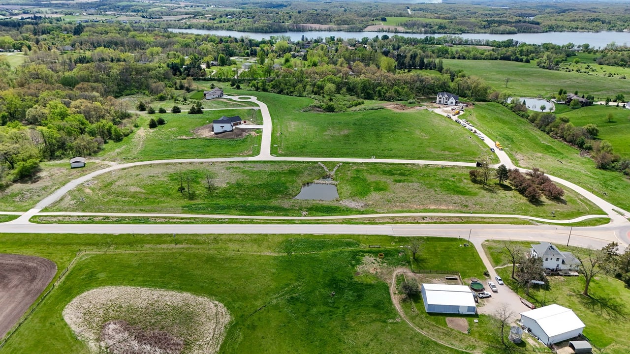 Aerial view of a rural development with winding roads, spacious green lots, scattered homes, and a small pond.