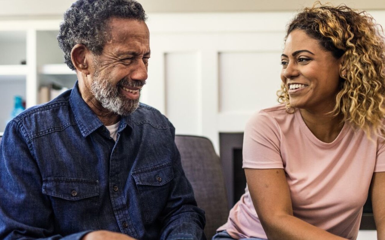 A smiling older man and woman sit together indoors, facing one another in conversation.