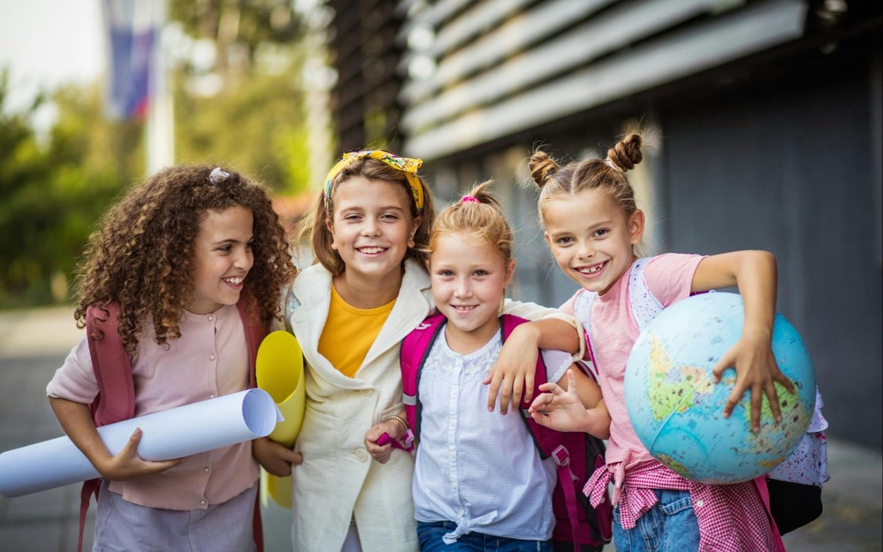 Four schoolgirls with backpacks pose together, one holding a globe and another carrying rolled papers.