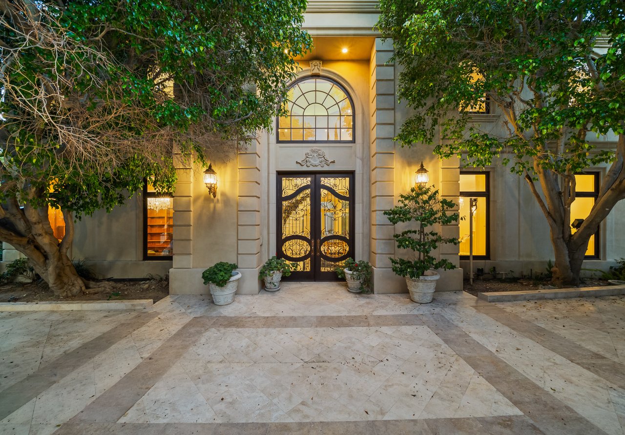 Grand stone home entrance with ornate iron doors, an arched window, and a tiled forecourt at night.