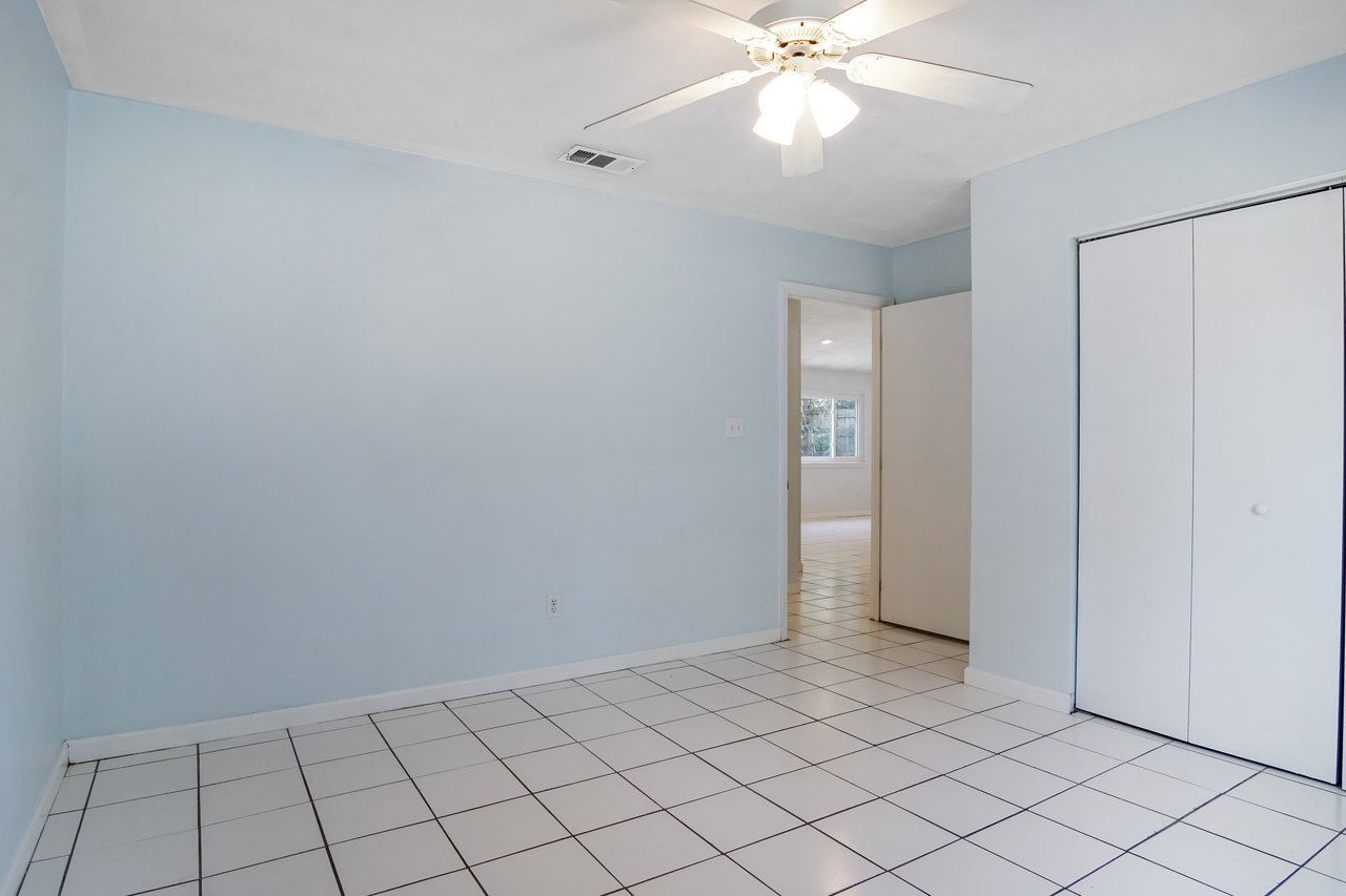 Empty room with pale blue walls, white tiled floor, and ceiling fan. An open door reveals another room with natural light coming through a window.