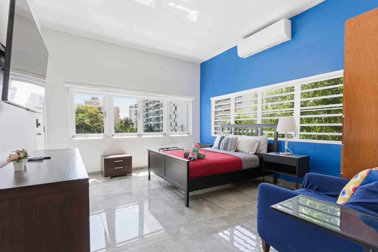 Furnished bedroom with a red-covered bed, bright blue accent wall, and windows overlooking greenery.