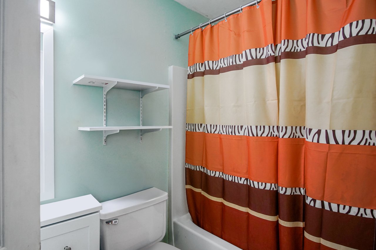Bathroom at 2506 Nugget Lane in Tallahassee, Florida, with light green walls and white fixtures. The space includes a toilet, built-in shelving, and a shower-tub combination with a colorful striped curtain in orange, beige, and brown tones.