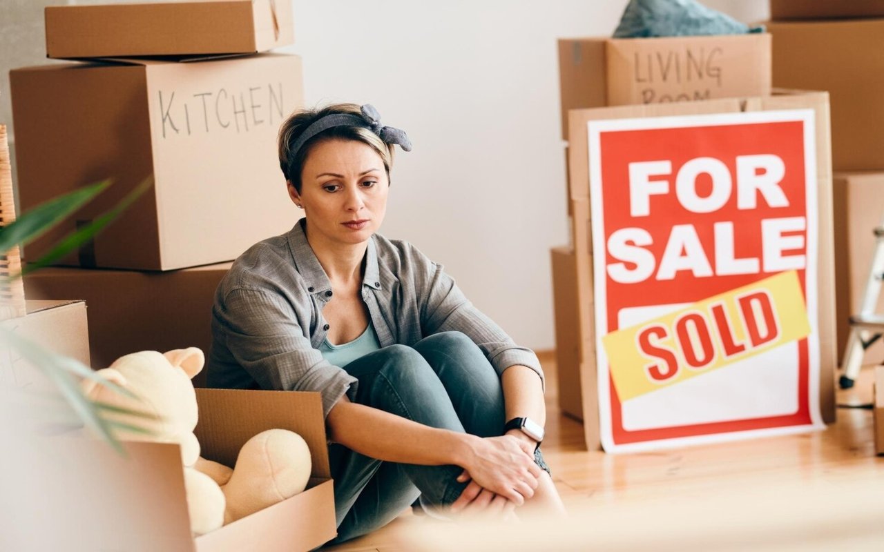 A woman sits among moving boxes beside a For Sale sign marked Sold.