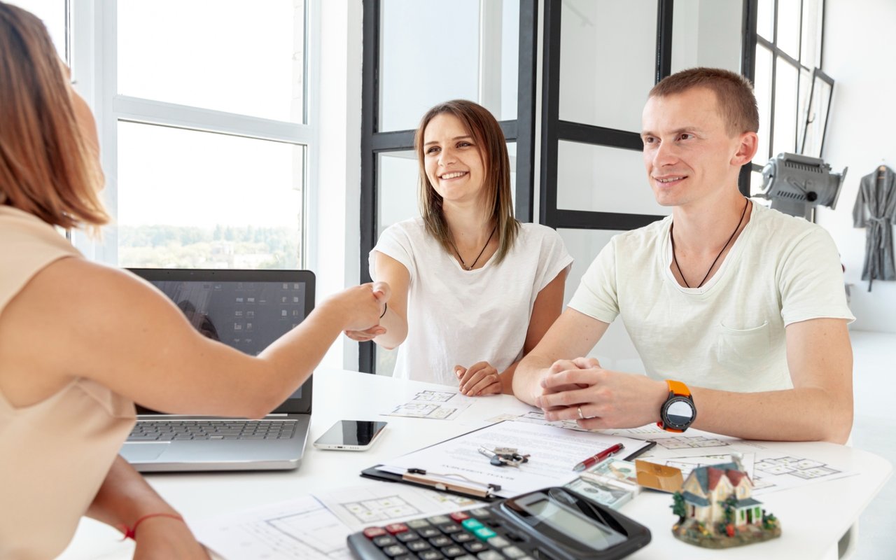 Real estate agent shakes hands with a smiling couple across a table of property plans.