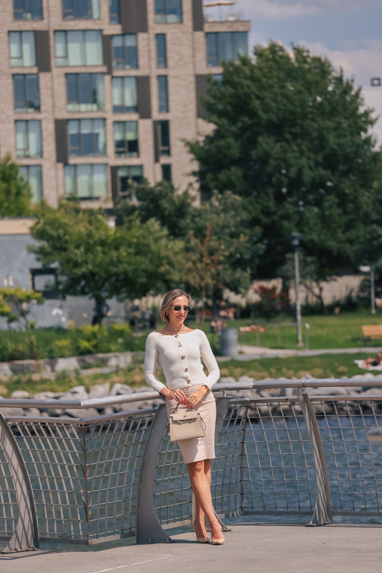 Woman in sunglasses standing on a waterfront bridge with a modern apartment building behind her.