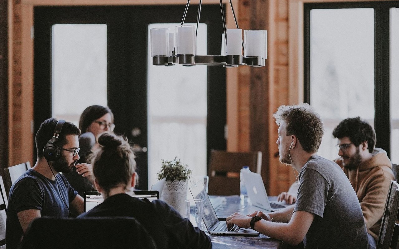 Young adults work on laptops around a table beneath a modern chandelier.