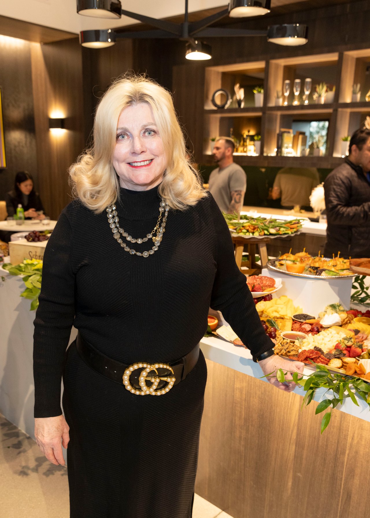 Blonde woman in black beside a buffet spread at an indoor reception.