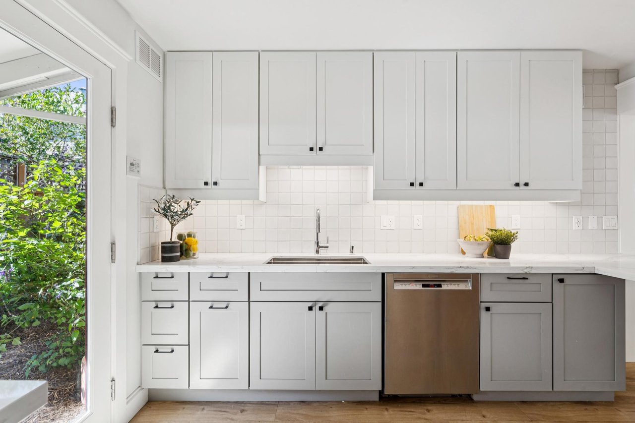 Bright kitchen with white cabinets, a tiled backsplash, and a glass door overlooking a leafy garden.