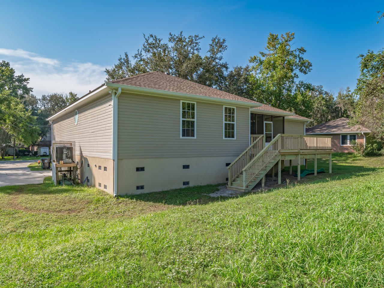 Rear of a raised pale green house with an elevated wooden deck and stairs above a sloping grassy yard.