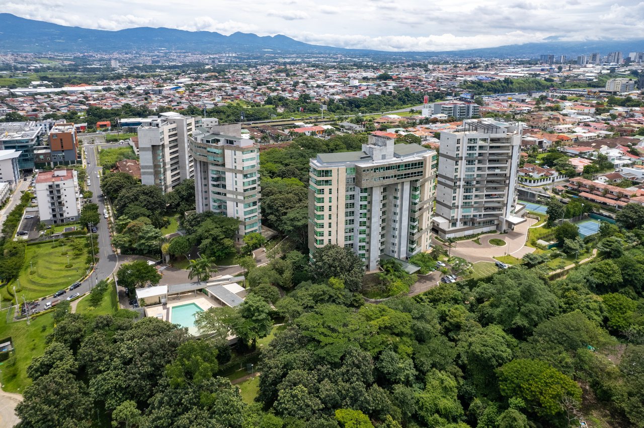 Aerial view of high-rise residences amid green neighborhoods, with a cityscape and distant mountains.