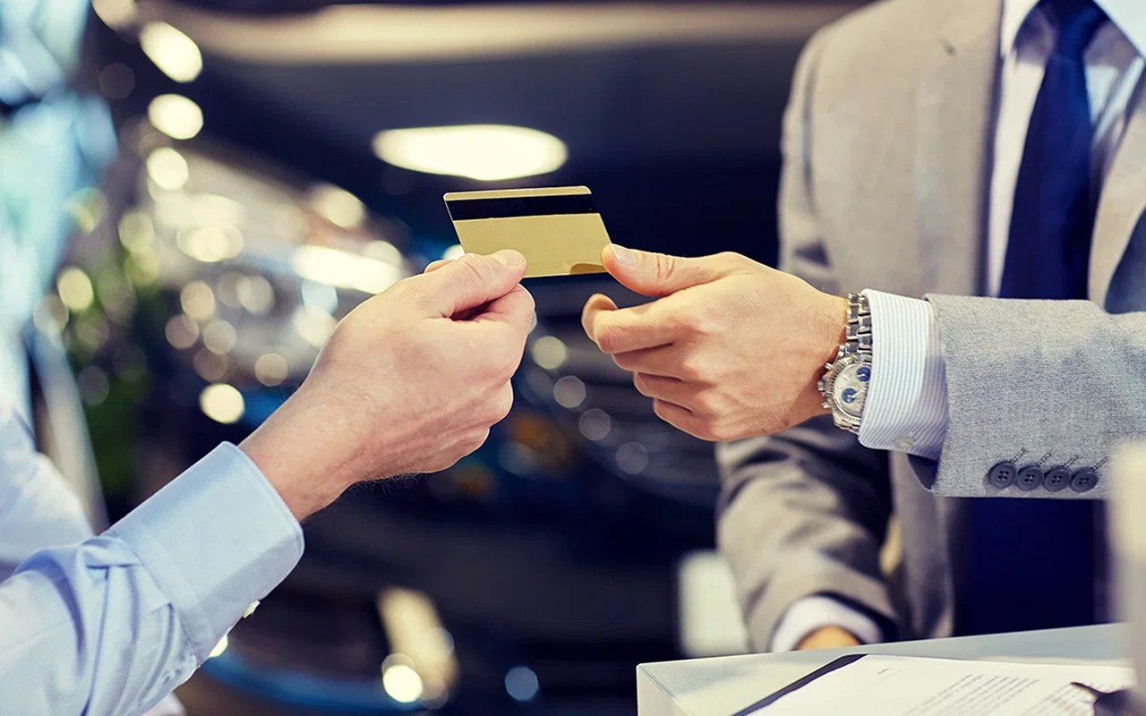 Two people exchange a gold-colored credit card across a counter; one wears a gray suit and wristwatch.