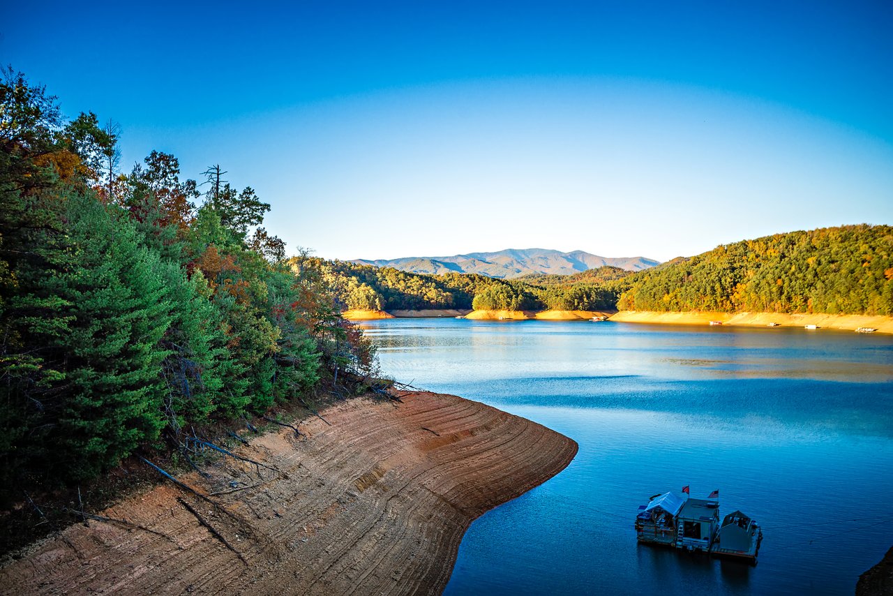 Fontana Lake in autumn with a wide band of exposed red clay shoreline below the summer waterline, a pontoon boat anchored in the cove and Smoky Mountain ridges beyond