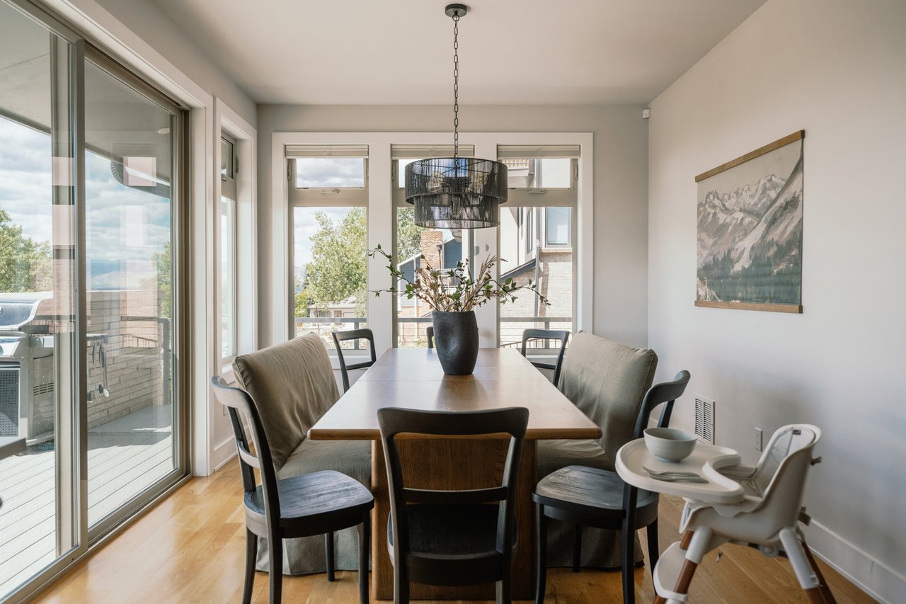 Dining room with a wooden table, upholstered chairs, and bright windows beside a balcony.