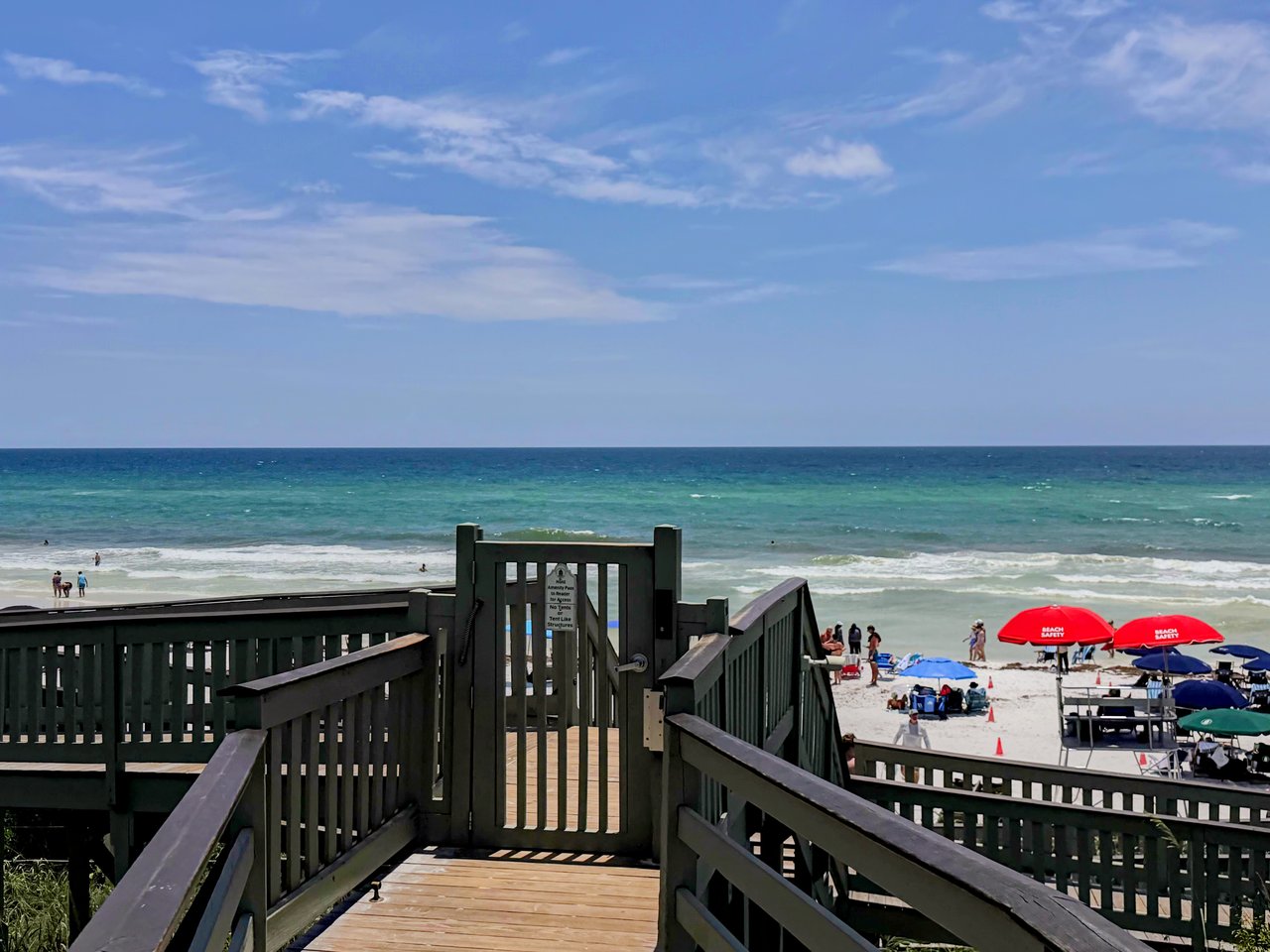 Sunny beach view of the emerald green waters and white sand in Rosemary Beach, Florida, featuring rows of green beach umbrellas and a wooden boardwalk along the Scenic Highway 30A coastline.
