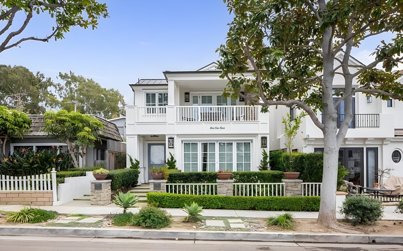 A large, modern, two-story white house with a balcony and a well-kept front yard with a white picket fence.