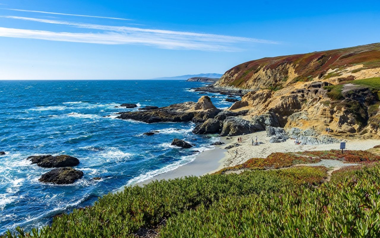 Rocky coastline and small sandy cove beneath rugged bluffs and a bright blue sky.