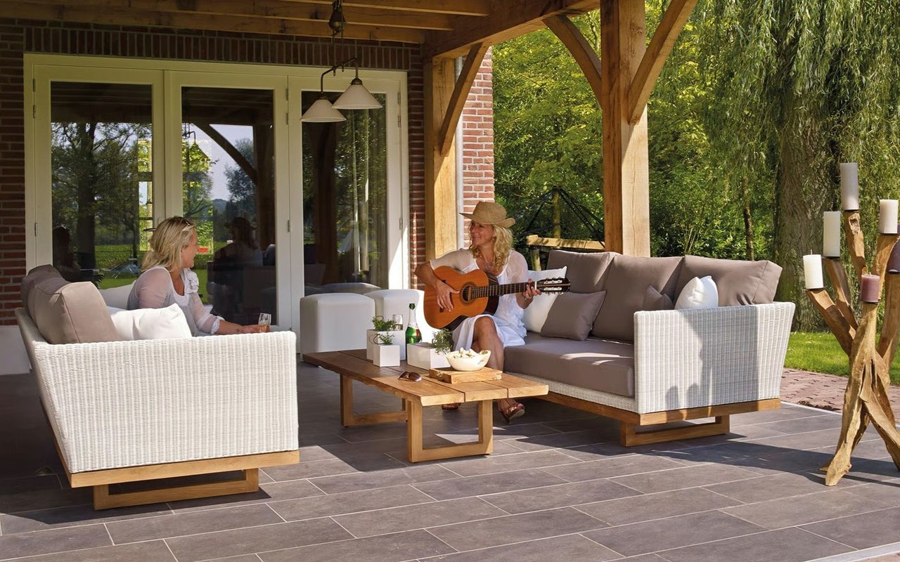 Covered patio with wicker seating and timber beams; two women relax as one plays guitar.