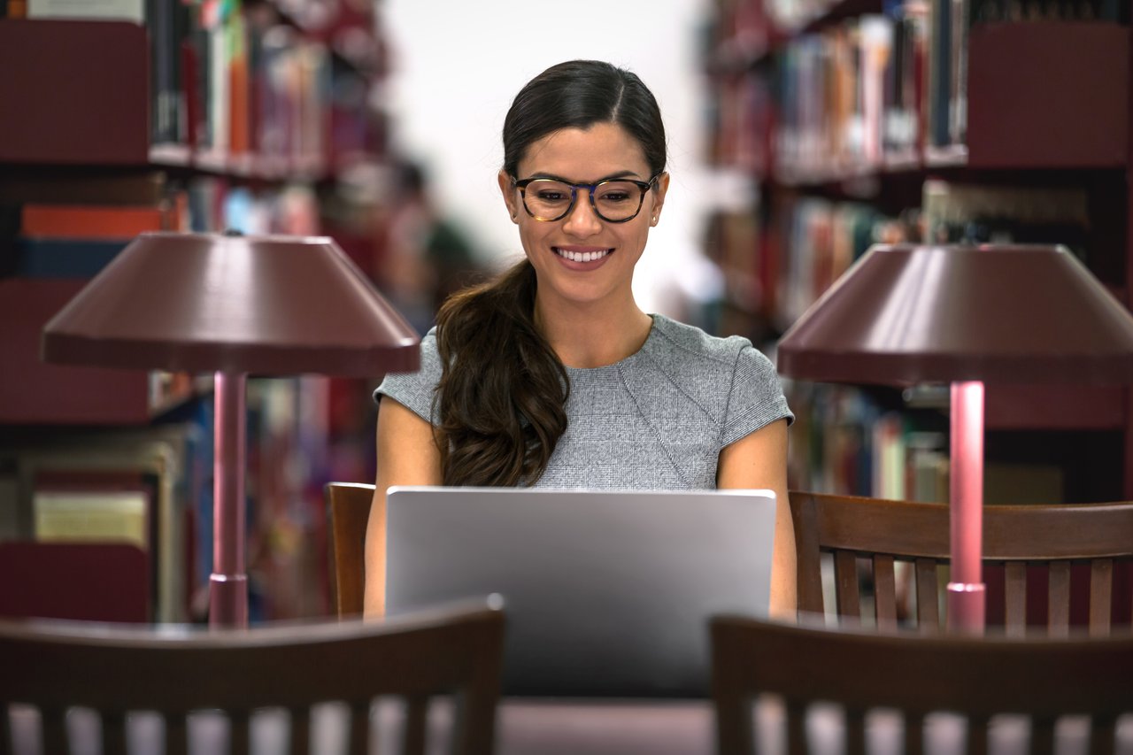 Woman studying for the real estate exam in a library, smiling while working on a laptop between two desk lamps, surrounded by bookshelves.