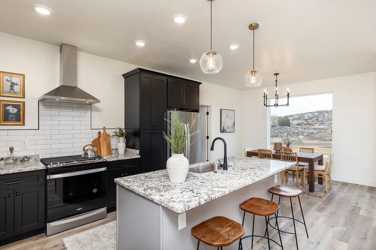 Kitchen with black cabinets, a granite island and bar stools, opening to a dining area beside a wide window.