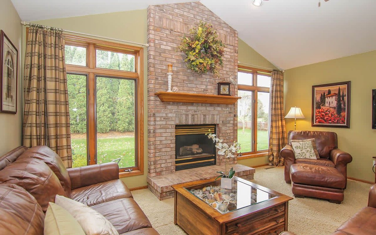 Living room with a tall brick fireplace, large yard-facing windows, and brown leather seating.