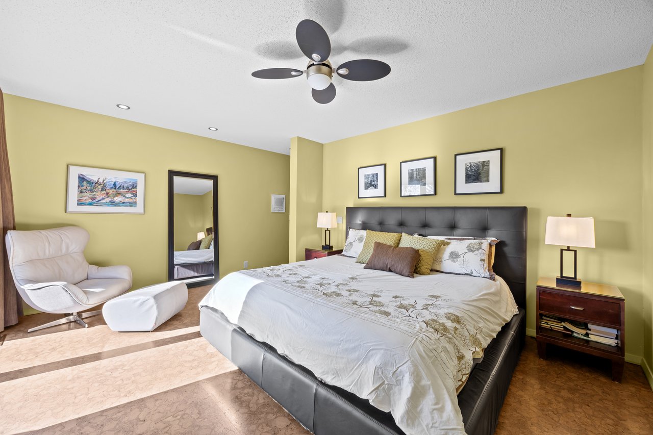 Furnished bedroom with a large dark bed, patterned bedding, ceiling fan, and white lounge chair.
