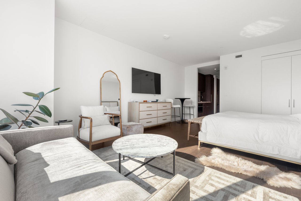 Furnished studio bedroom and living area with a white bed, gray sofa, and round coffee table.