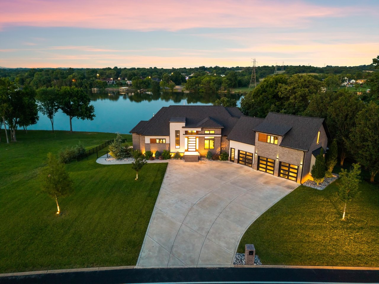 Aerial view of a modern lakeside home with a broad curved driveway and illuminated windows at dusk.