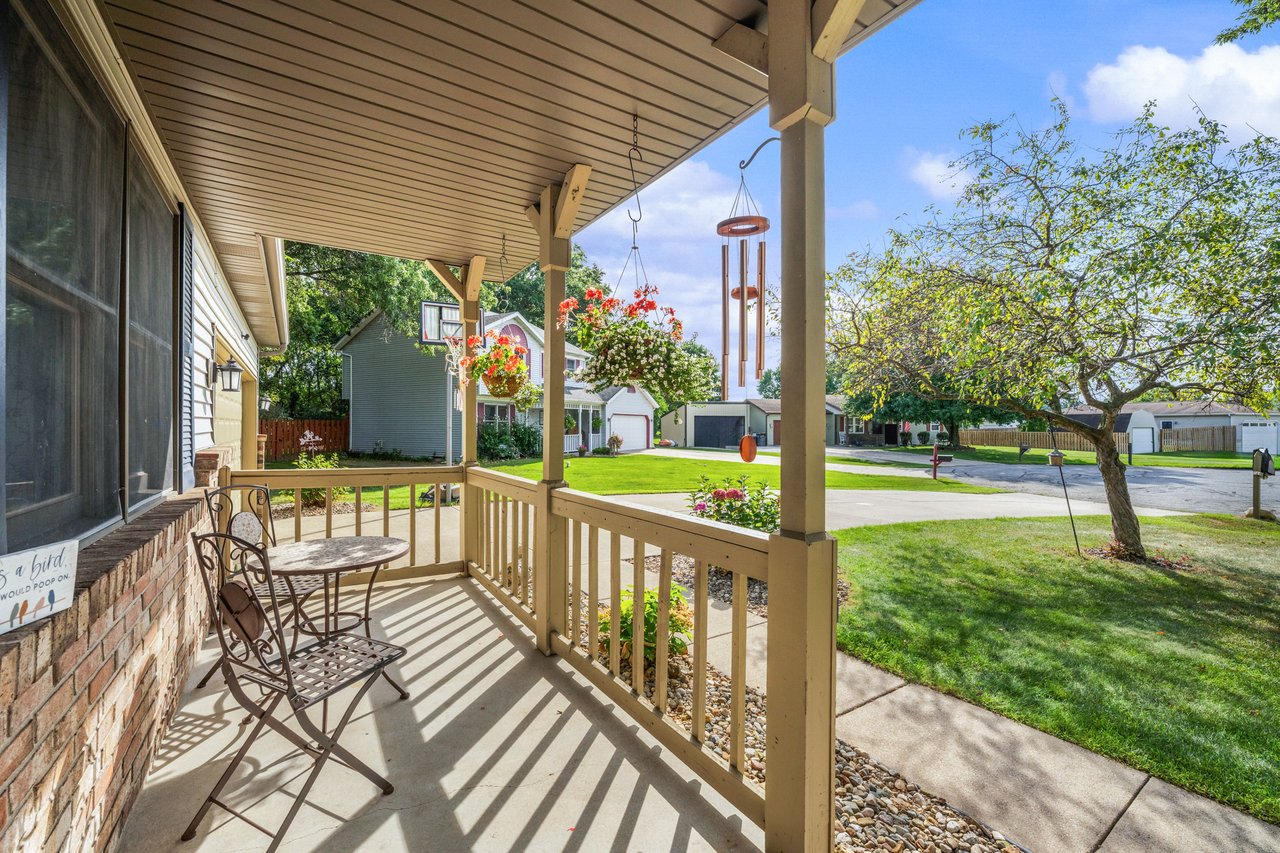 Covered front porch with metal seating, hanging flowers, and a view of the sunny lawn and neighboring homes.
