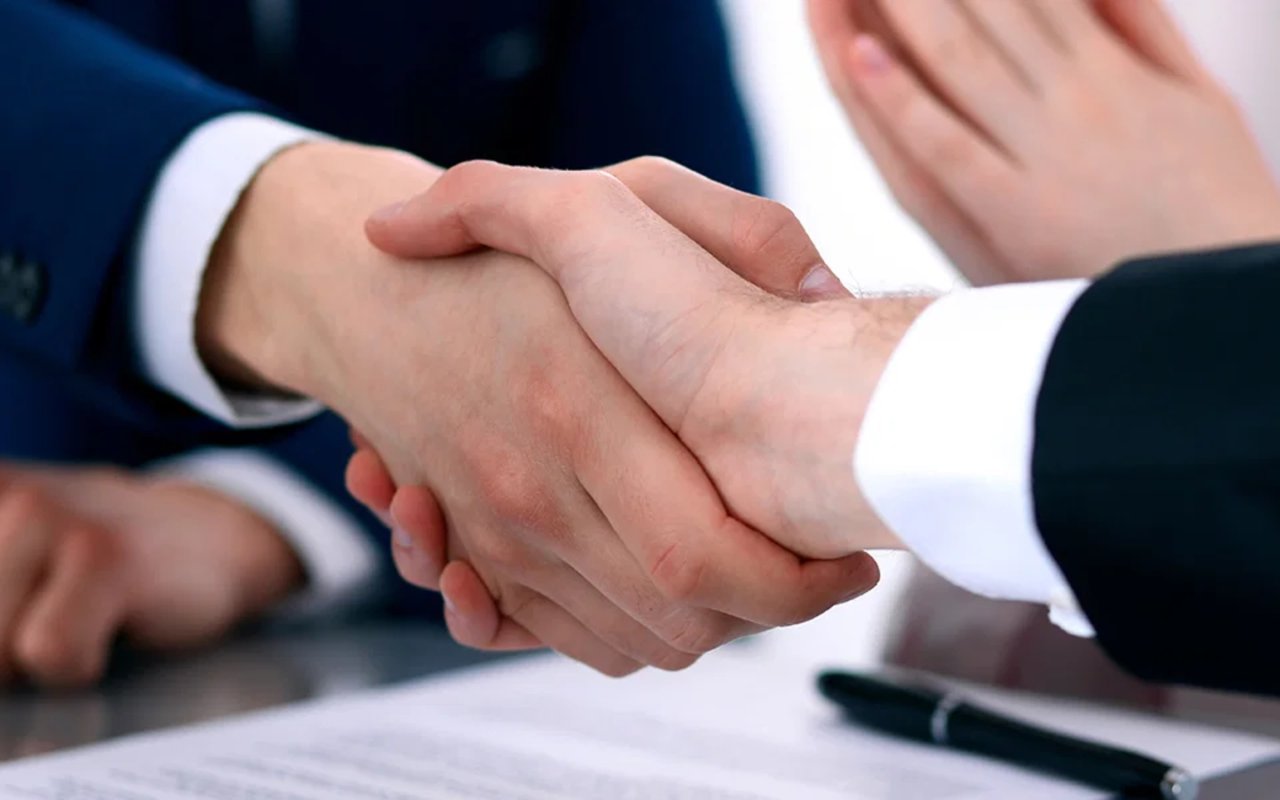 Two people in business suits shake hands over paperwork on a desk.