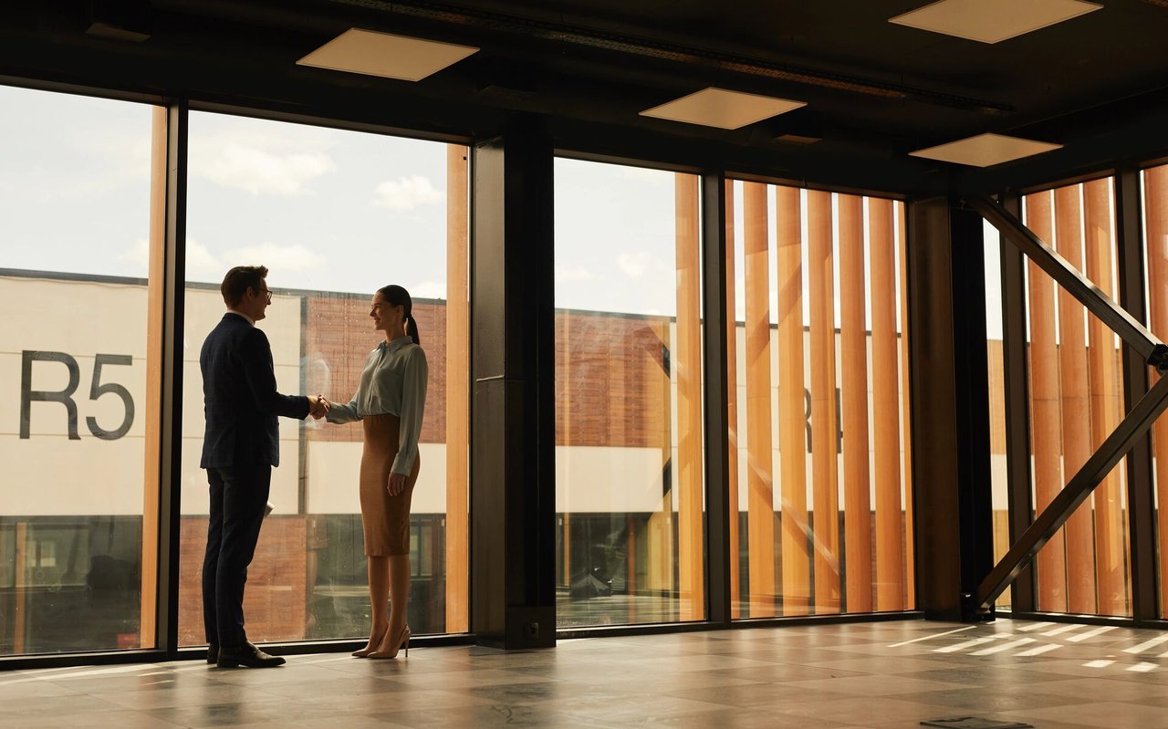 Two people shake hands beside floor-to-ceiling windows in a modern office.