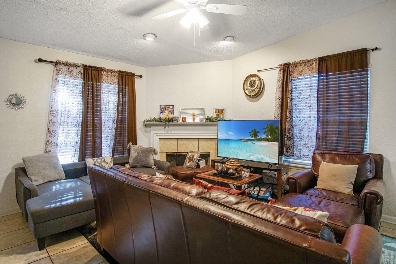 Living room at 2506 Nugget Lane in Tallahassee, Florida. The space features a tile floor, brown leather sofa and armchair, gray sectional, and a wall-mounted fireplace with white mantel. A flat-screen TV sits to the right of the fireplace, with two windows dressed in patterned curtains bringing in natural light.