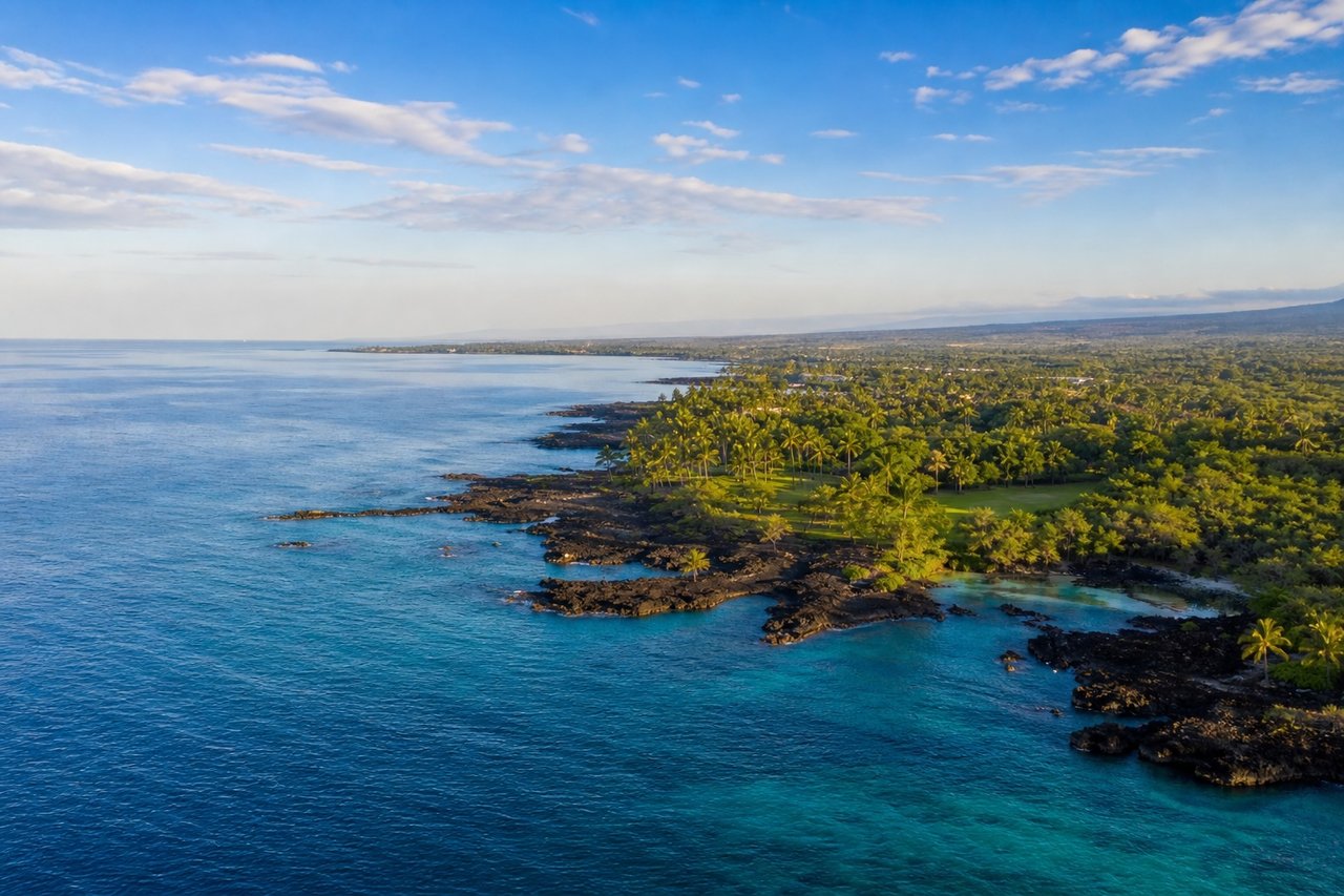 Aerial view of Hawaii’s Kona coastline with calm turquoise water, black lava rock shoreline, lush green vegetation, and a bright blue sky with light clouds.