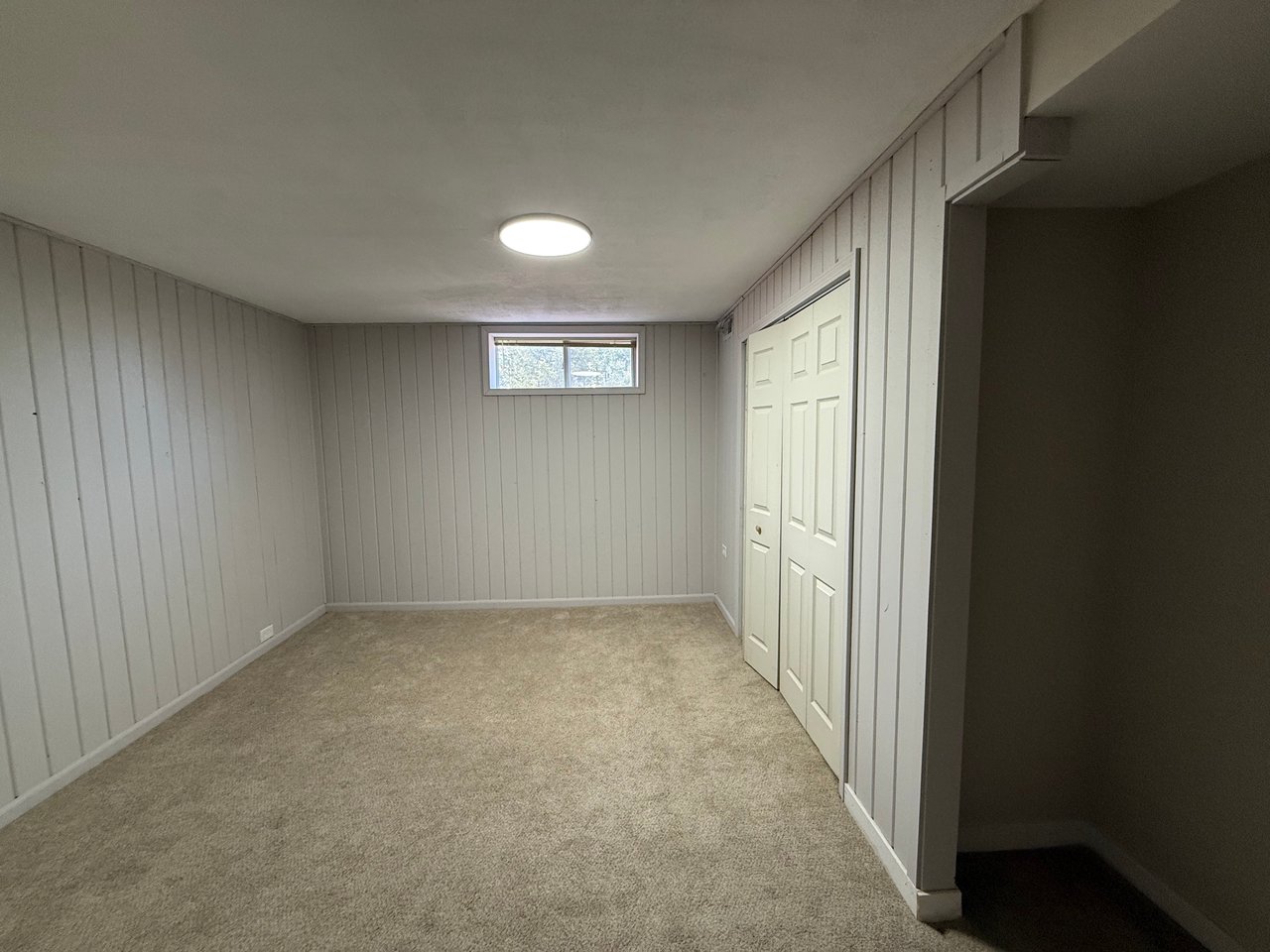 Empty basement room with beige carpet, paneled walls, a small high window, and folding closet doors.
