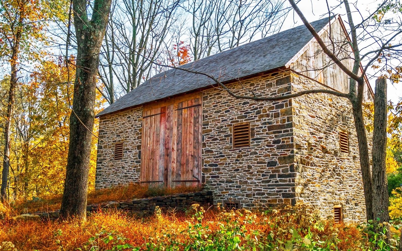 Stone barn with weathered wooden doors surrounded by autumn trees.