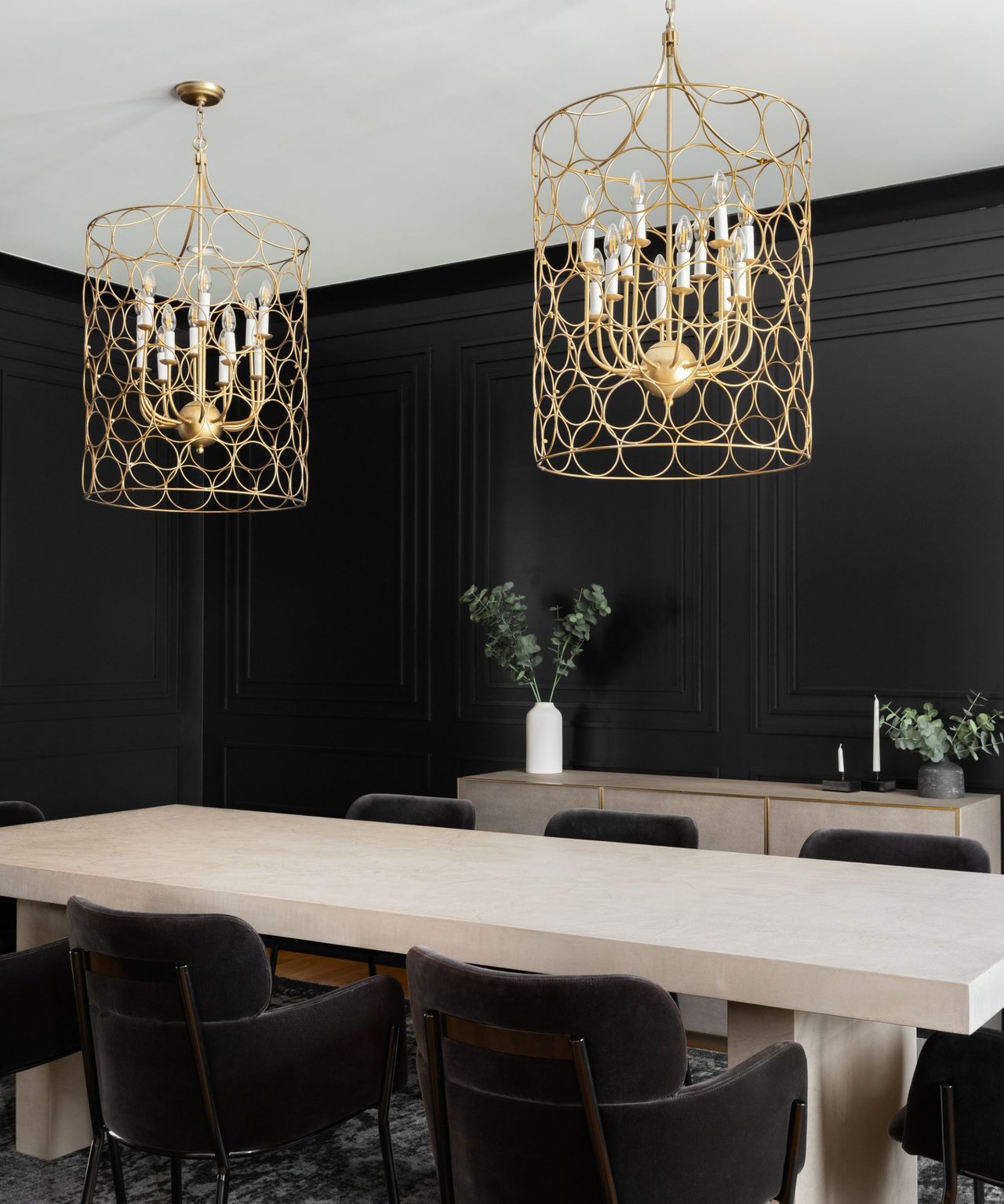 Dining room with a pale stone table, dark chairs, black paneled walls, and gold cage chandeliers.