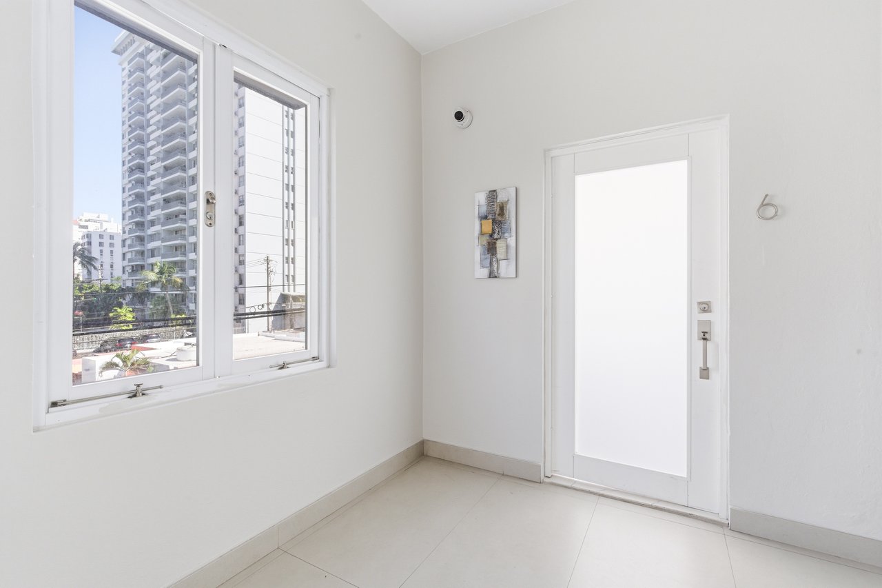 Vacant white room with a large window overlooking nearby apartment buildings and a white door.