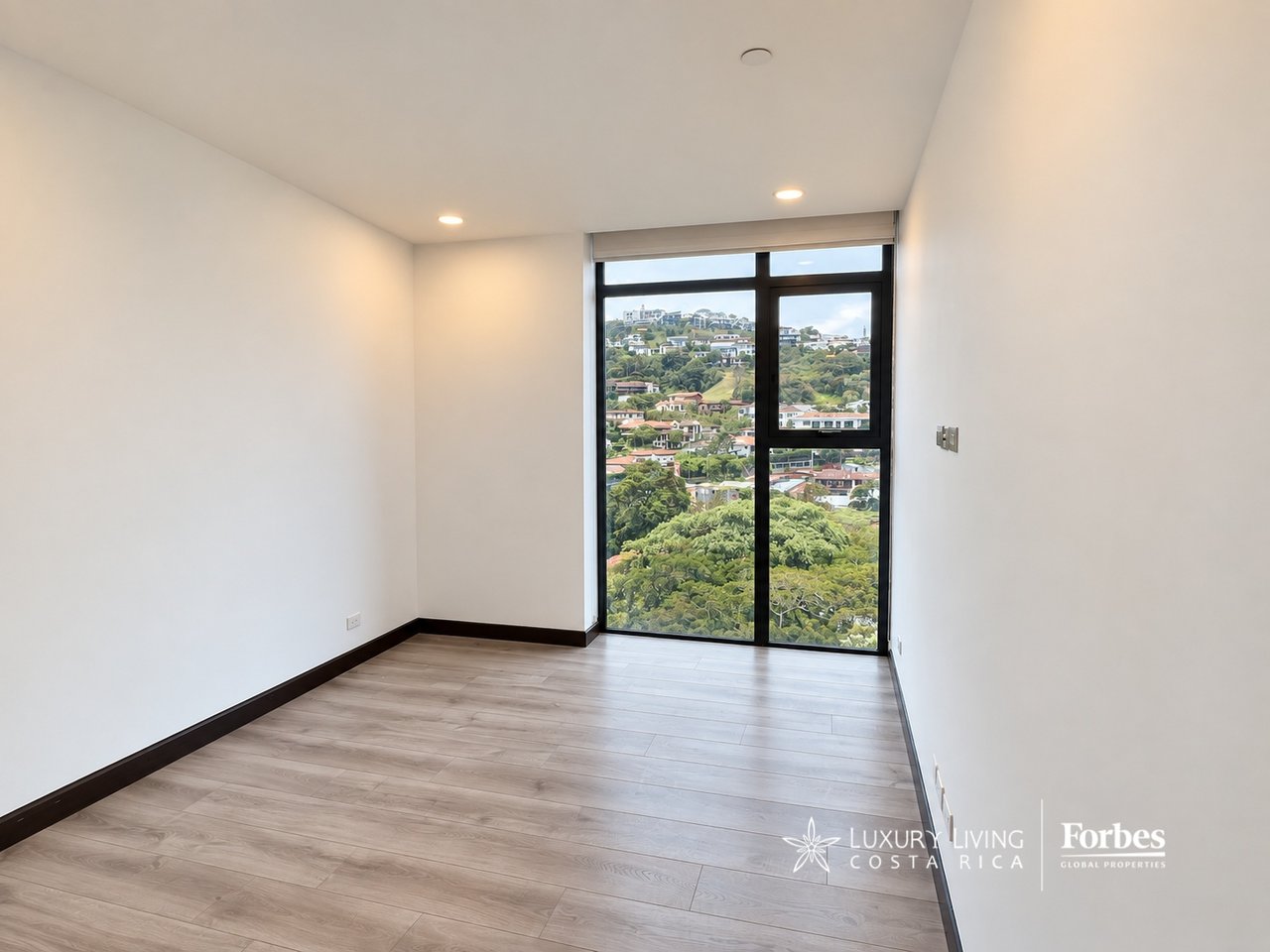 Empty apartment room with wood flooring and a large window overlooking green hills and homes.