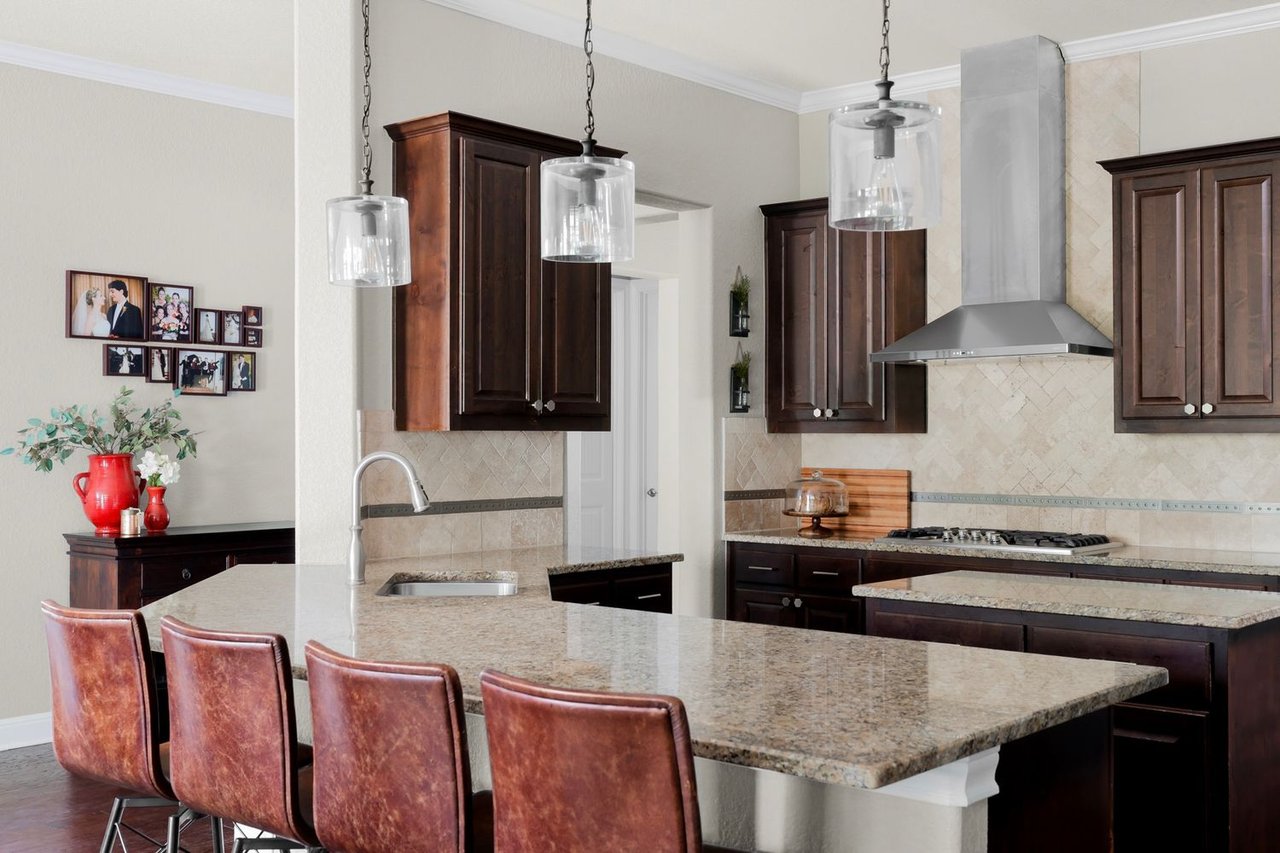 Kitchen with dark wood cabinets, granite island, leather bar stools, and stainless steel range hood.