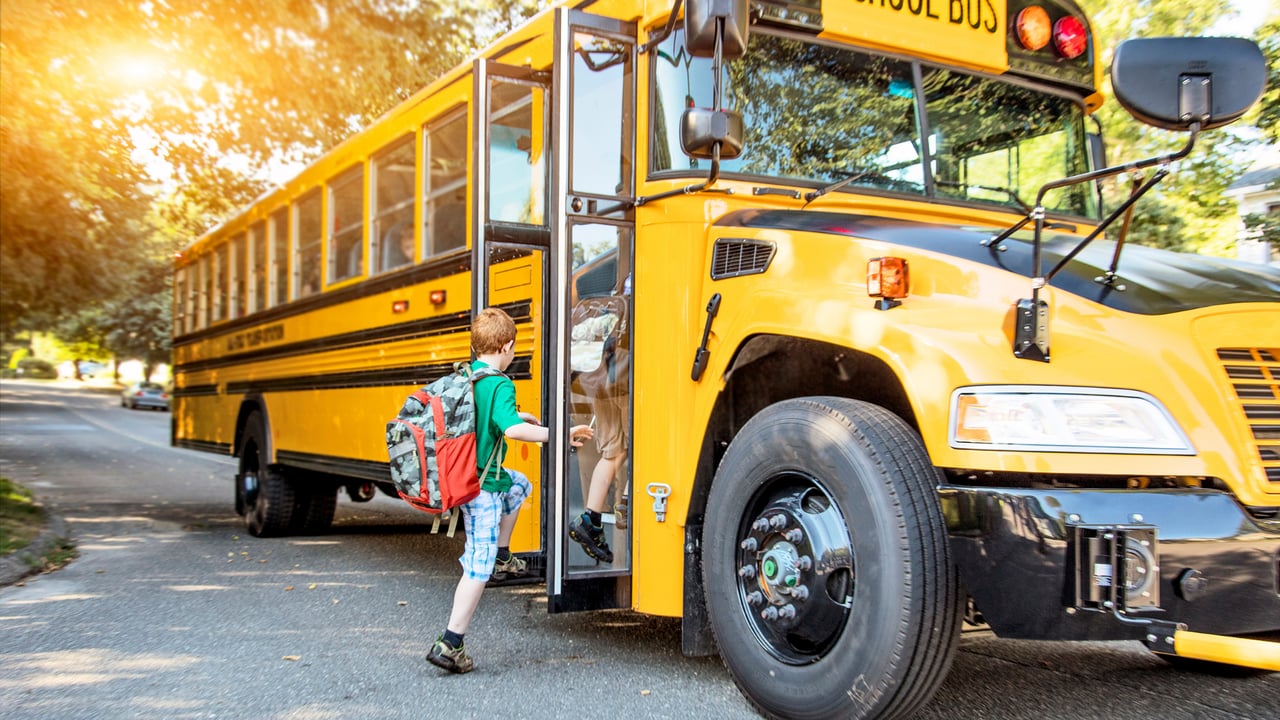 A child with a backpack boards a yellow school bus on a tree-lined street.