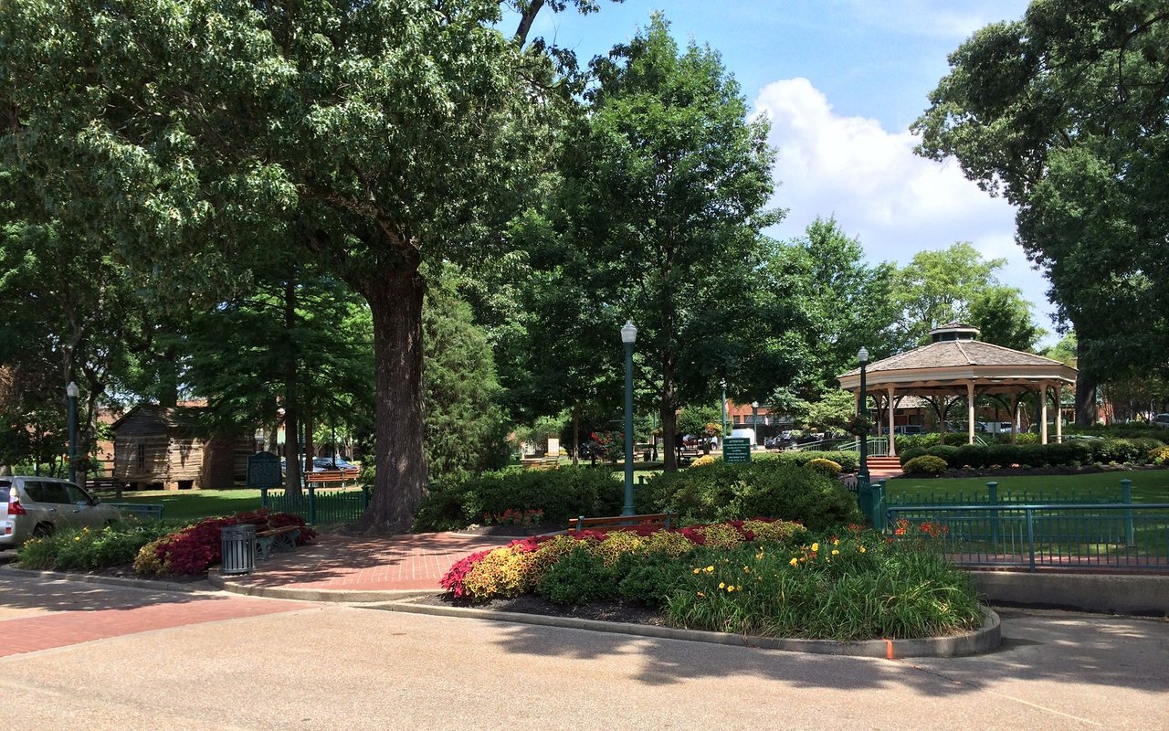 Shaded park with a wooden gazebo, brick walkways, and colorful flower beds.