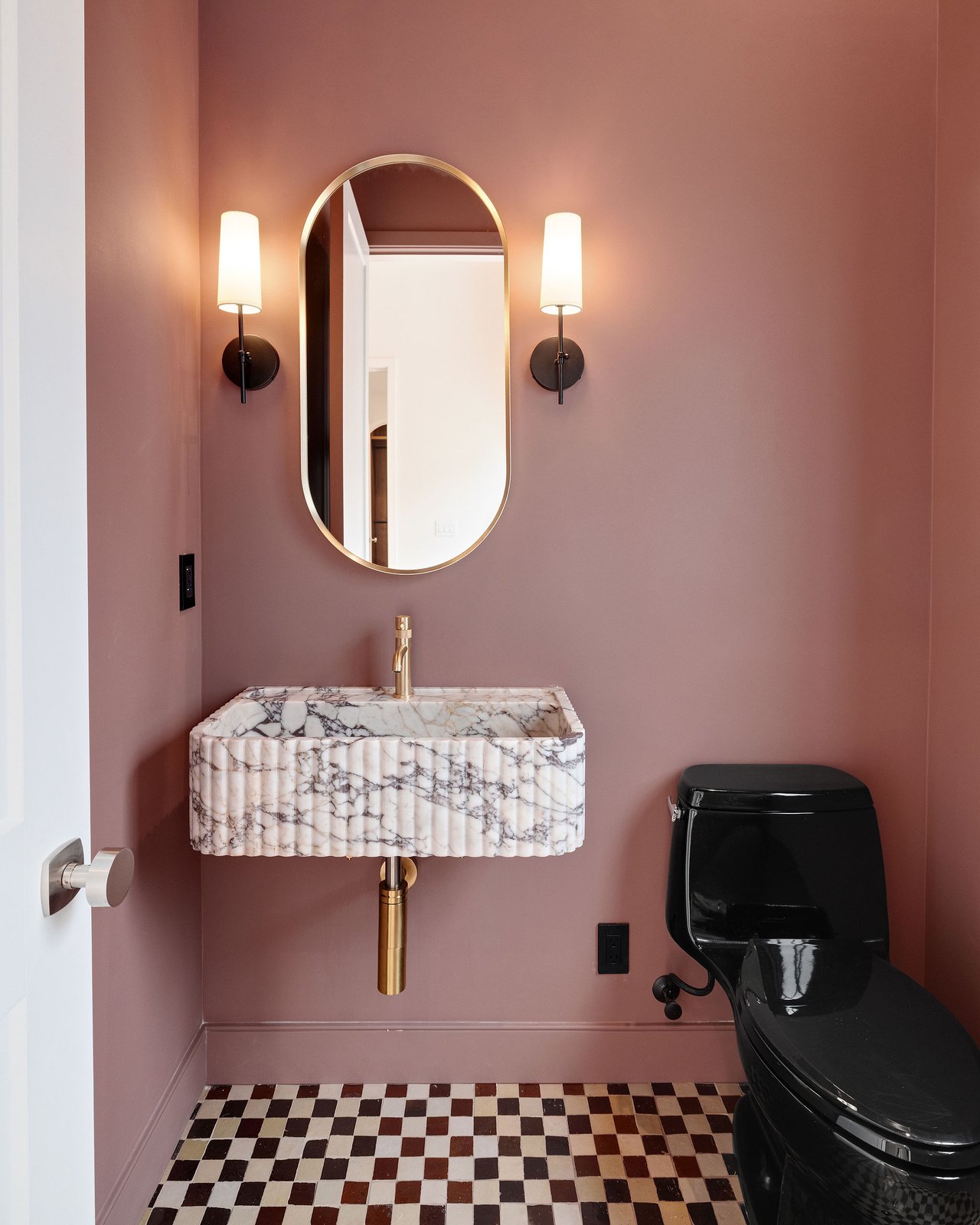 Powder room with a fluted marble vanity, brass fixtures, rose walls, and checkerboard floor tiles.