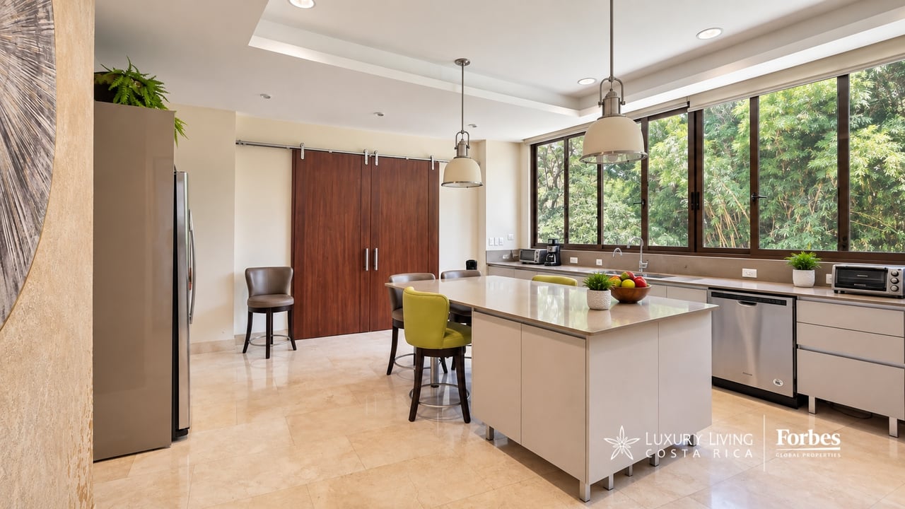Modern kitchen with a large island, pendant lights, and wide windows overlooking greenery.